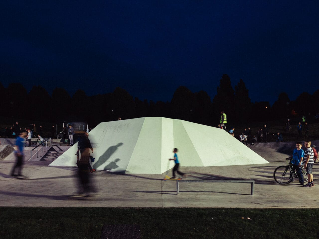 A white wheels park surrounded by people standing and people riding bikes.