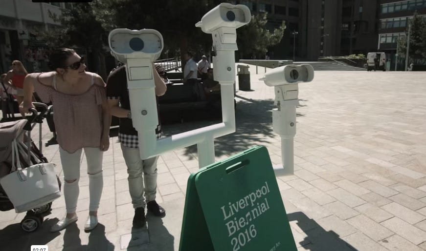 An installation which painted white tubular metal pipes with three telescopes at varying heights. The installation is within a city centre with people walking around