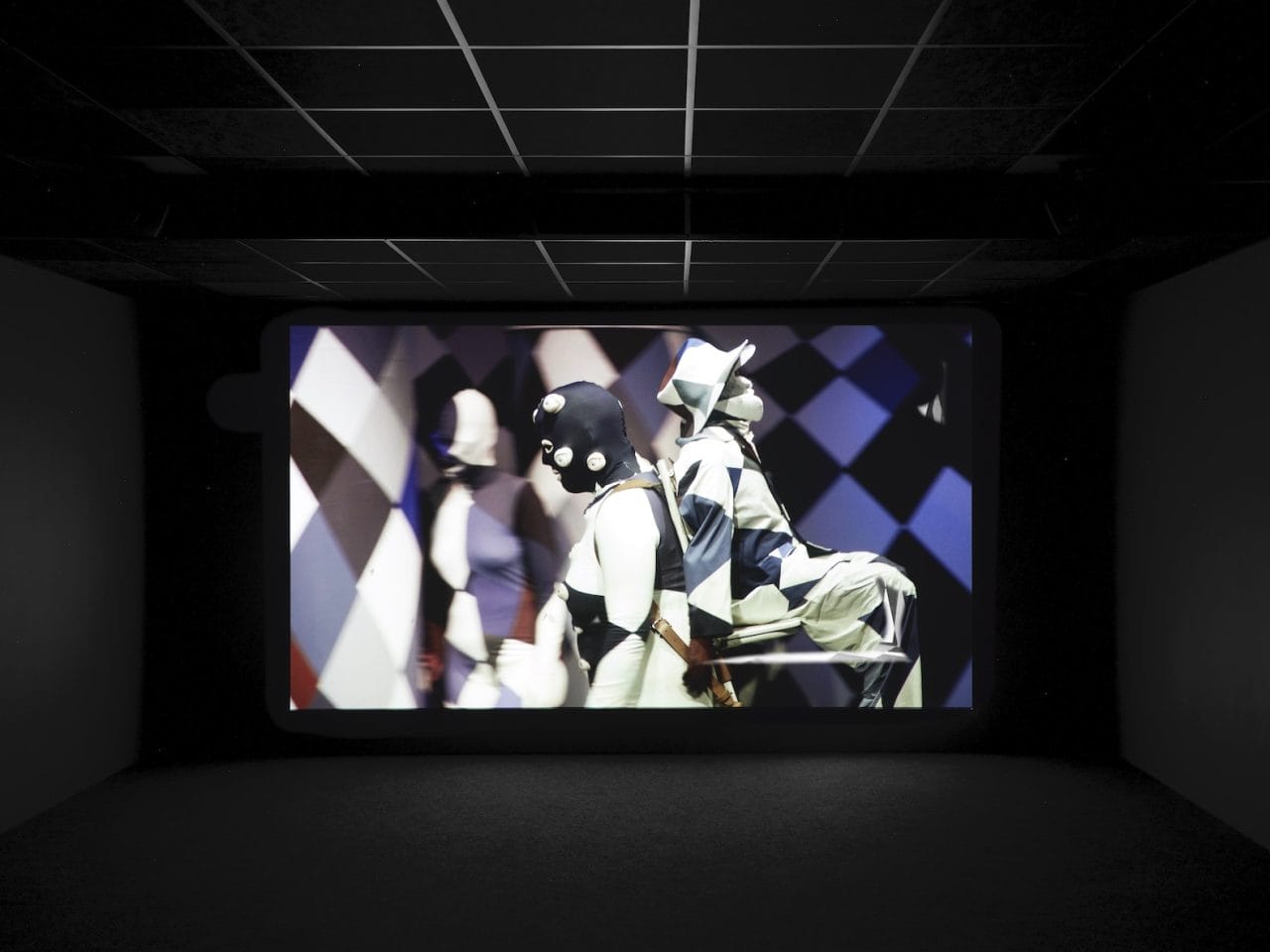 Someone walking, with a person sat on a chair strapped to their back. The people and the room they are in are all camouflaged together in a blue and white checkered pattern.