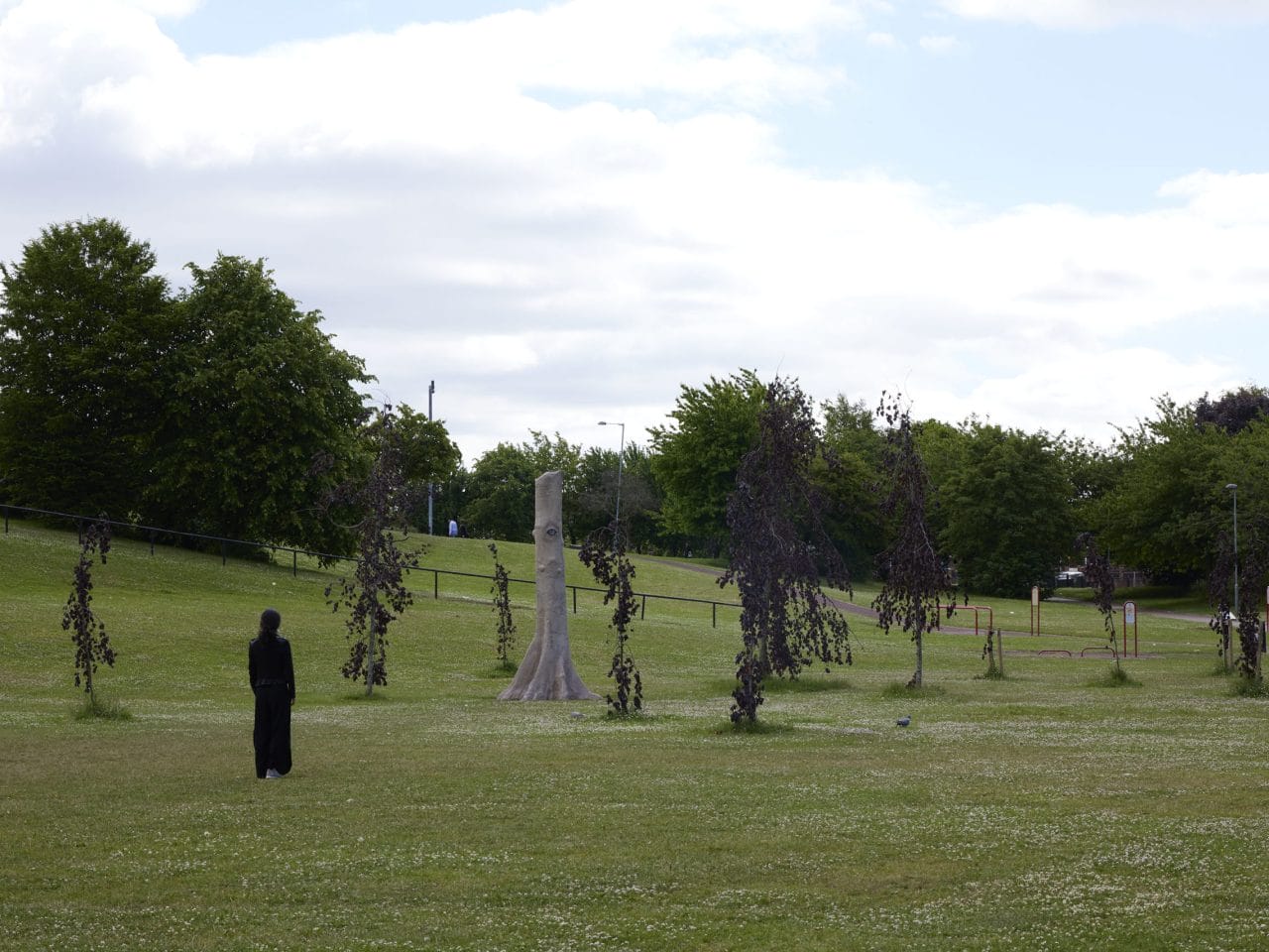 A sculpture amongst some trees on a feild, the sculpture is made to disguise as a tree and has a realistic orange/brown eye.