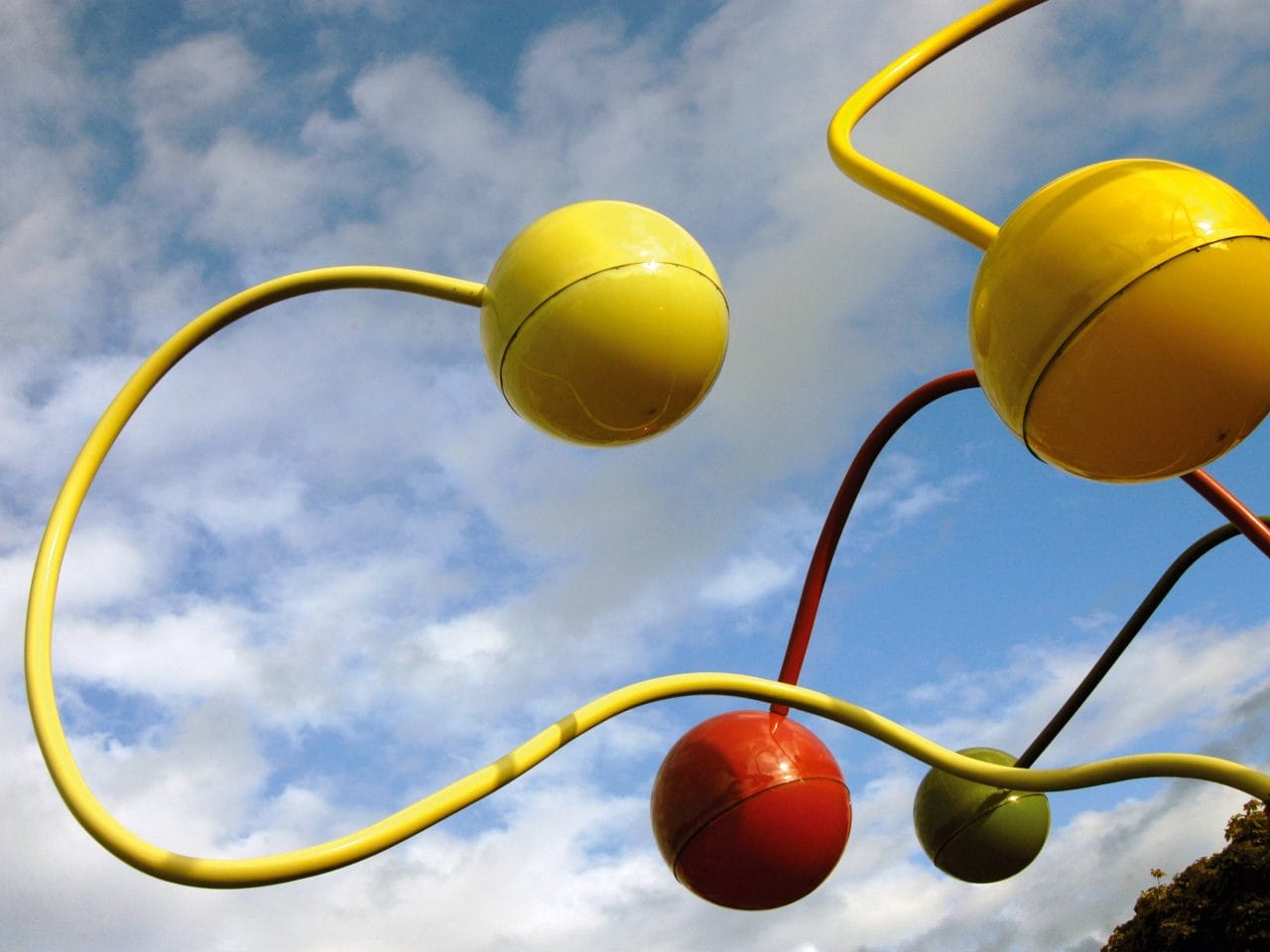 An outdoor artwork with red, yellow and green bendy poles that scale up through the trees and have a large ball at the end of it.
