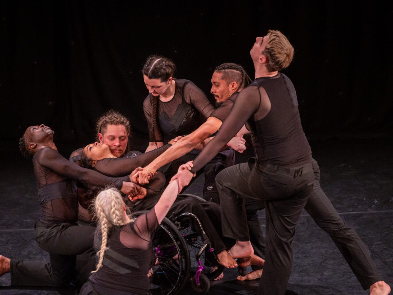 All seven dancers are connected holding hands. Arms are overlapped, some are stood up, some on the floor and in-between, all looking different ways. The wheelchair user is in the centre falling backwards into into another dancers.