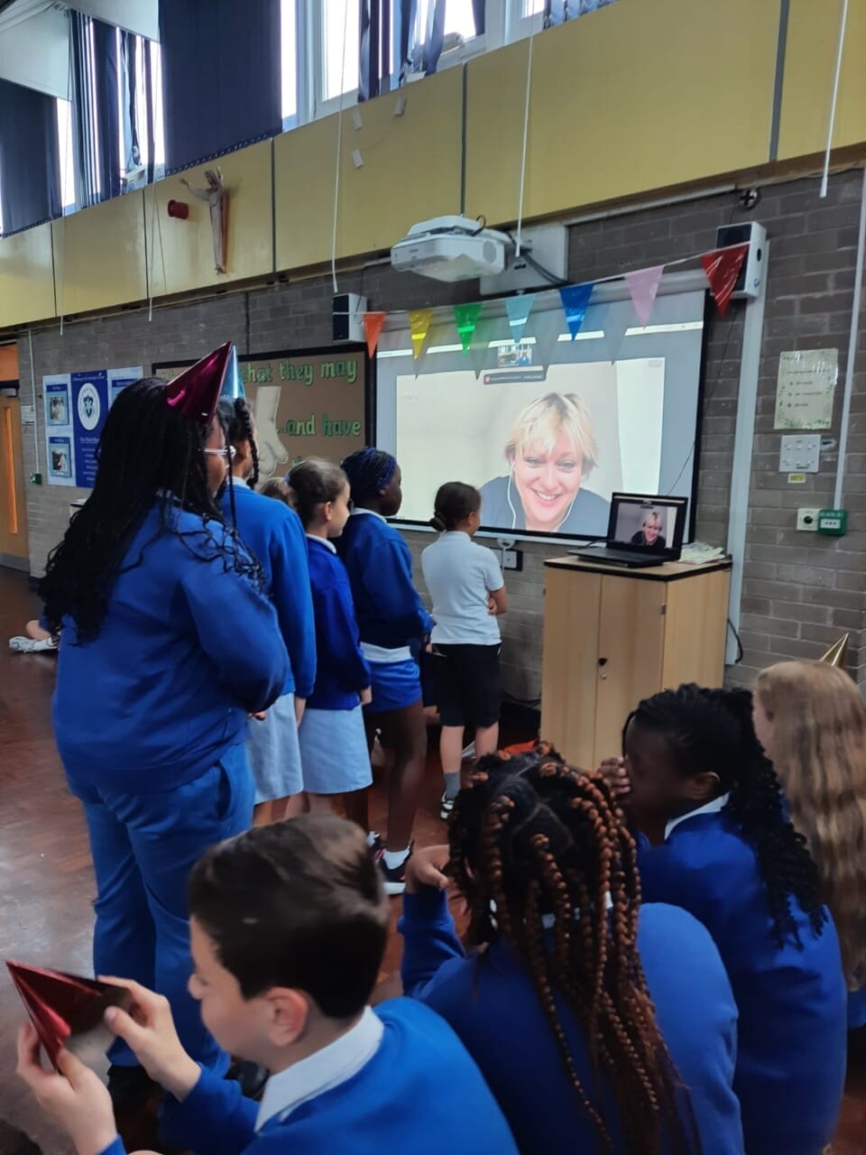 Children lining up wearing party hats, waiting to ask the artist a question as the artist is on zoom and projected on a screen