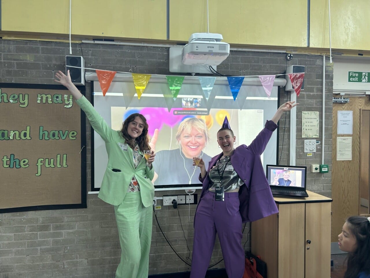 Two Liverpool Biennial Staff members standing in front of a projection screen wearing purple and green suits with matching party hats. On the projection is the artist Stine smiling with a balloon animation surrounding them.