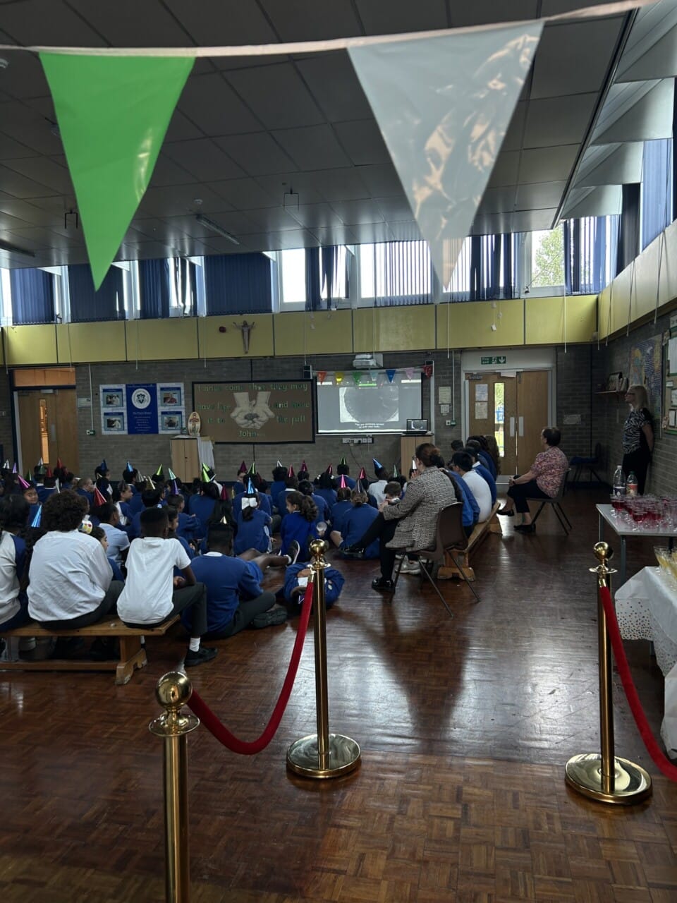 Bunting hanging from the top of the enterence and red ropes on golden stands as you wnter a room full of children sitting down on a school floor watching a film on a projection screen, they are all wearing party hats.