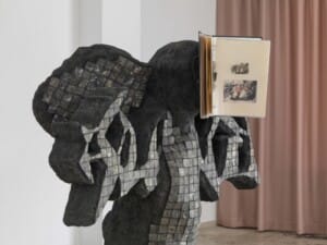 A ghost-like mosaic grey figure sculpture, there is a folder on the head section of the figure with plastic wallets inside which show smaller 2-D artworks.