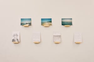 Seven artworks on a white wall held up with small wooden stands. The top line shows three seascape photographs and the bottom line shows two pencil drawings with two pieces of writing.