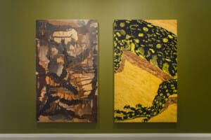 Two 2D artworks created with wax, on a dark green wall. The first artwork is a brown abstract piece and the second is detail of a reptile's body with a bright yellow background.