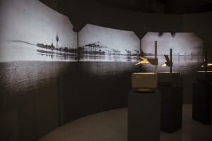 Three overhead projectors facing a wall, projected on the curved wall are black and white stills of the sea with scaffolding in the distance.