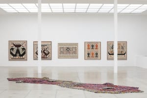 Five square artworks on a white gallery wall, they have repetitive patterns and symmetrical shapes on them. In the foreground there is a fabric work on the floor.