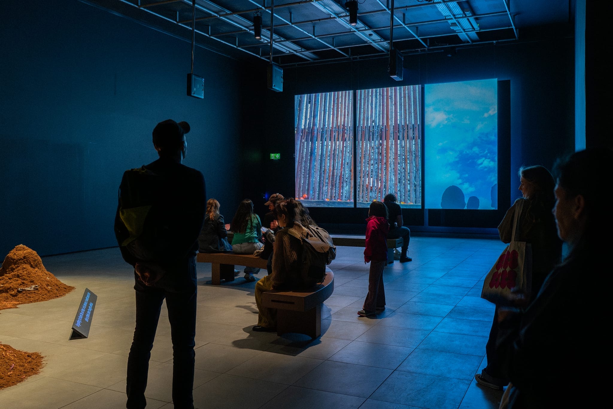 a selection of people in a dark gallery space with a large 3 screen projection and a mixed media installation behind them